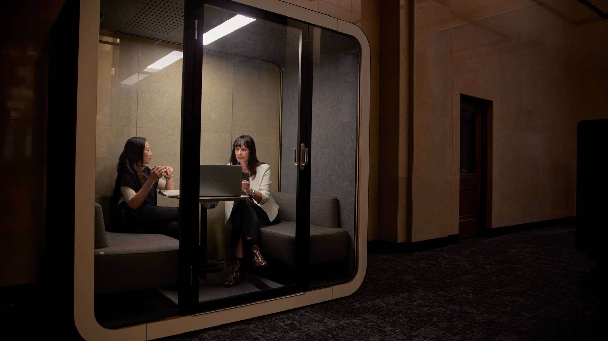 Two women are discussing in a meeting cube with a laptop on the desk