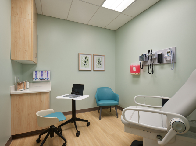 A clean, modern medical examination room with a laptop on a movable desk, a teal chair, an examination bed, wall-mounted medical tools, and minimal decor.