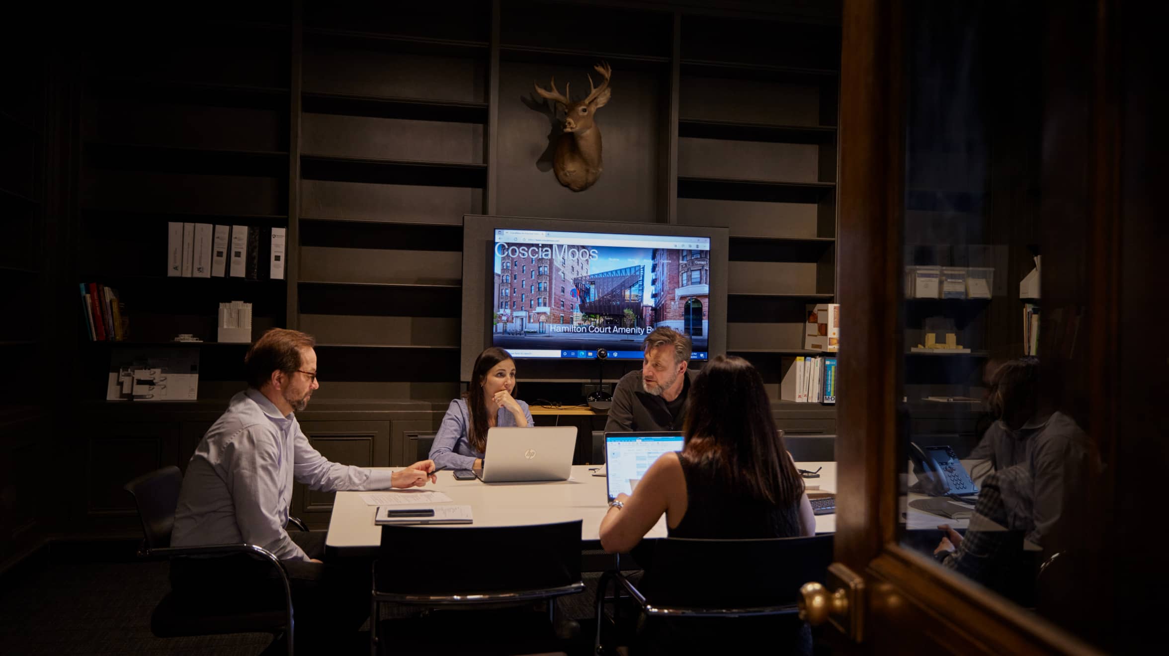 People in the meeting room are having a meeting with laptops on the desk, and a big screen behind them is displaying the CoscisMoos website.