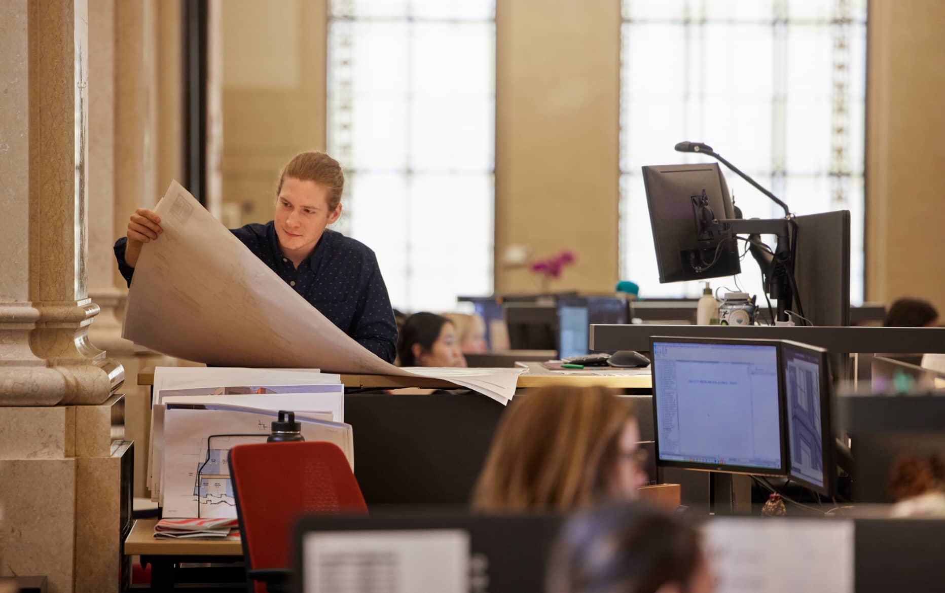 A man is standing at a standing desk, looking at blueprints, while others are seated and looking at an own screen.