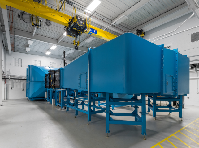 Large blue industrial machinery on platforms in a clean, well-lit factory setting with overhead yellow cranes.