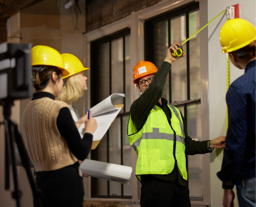 A man in an orange safety helmet is measuring a wall with a tape measure, while three others in yellow safety helmets are observing him and taking notes