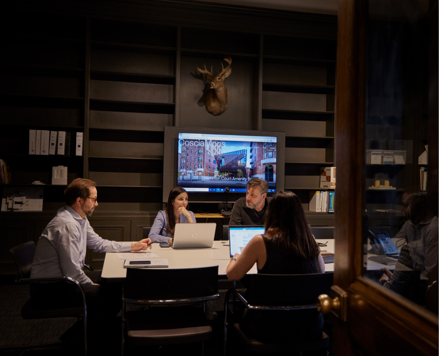 People in the meeting room are having a meeting with laptops on the desk, and a big screen behind them is displaying the CoscisMoos website.
